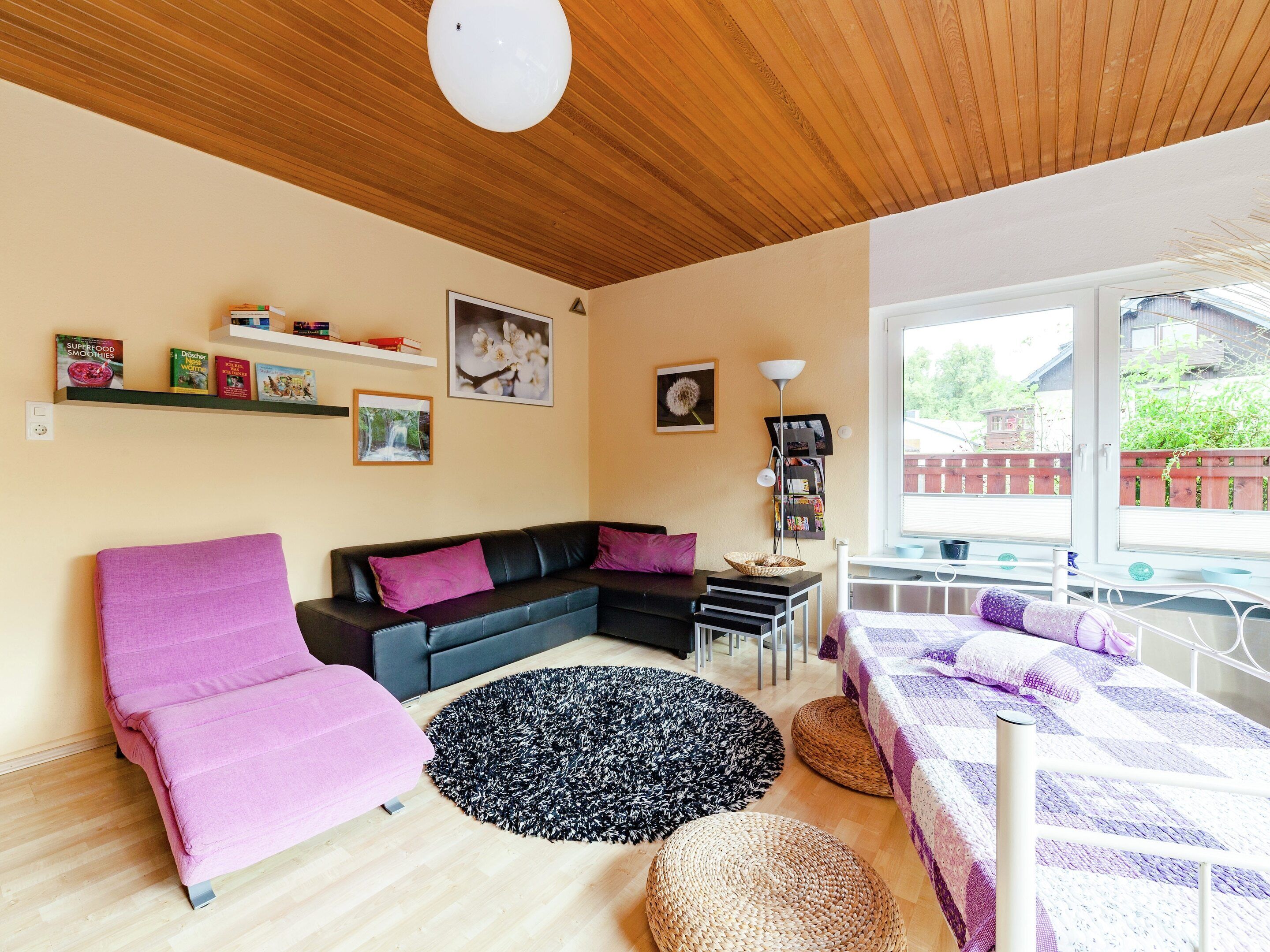 Una sala moderna con techo de madera, un sofá modular negro, un sillón rosa, un tapete redondo negro y una cama con manta a cuadros morada y blanca.