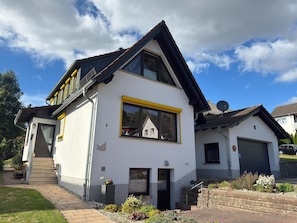 Une maison à deux étages avec un toit en pente, une grande fenêtre ornée d’un cadre jaune et une antenne parabolique sur le toit.