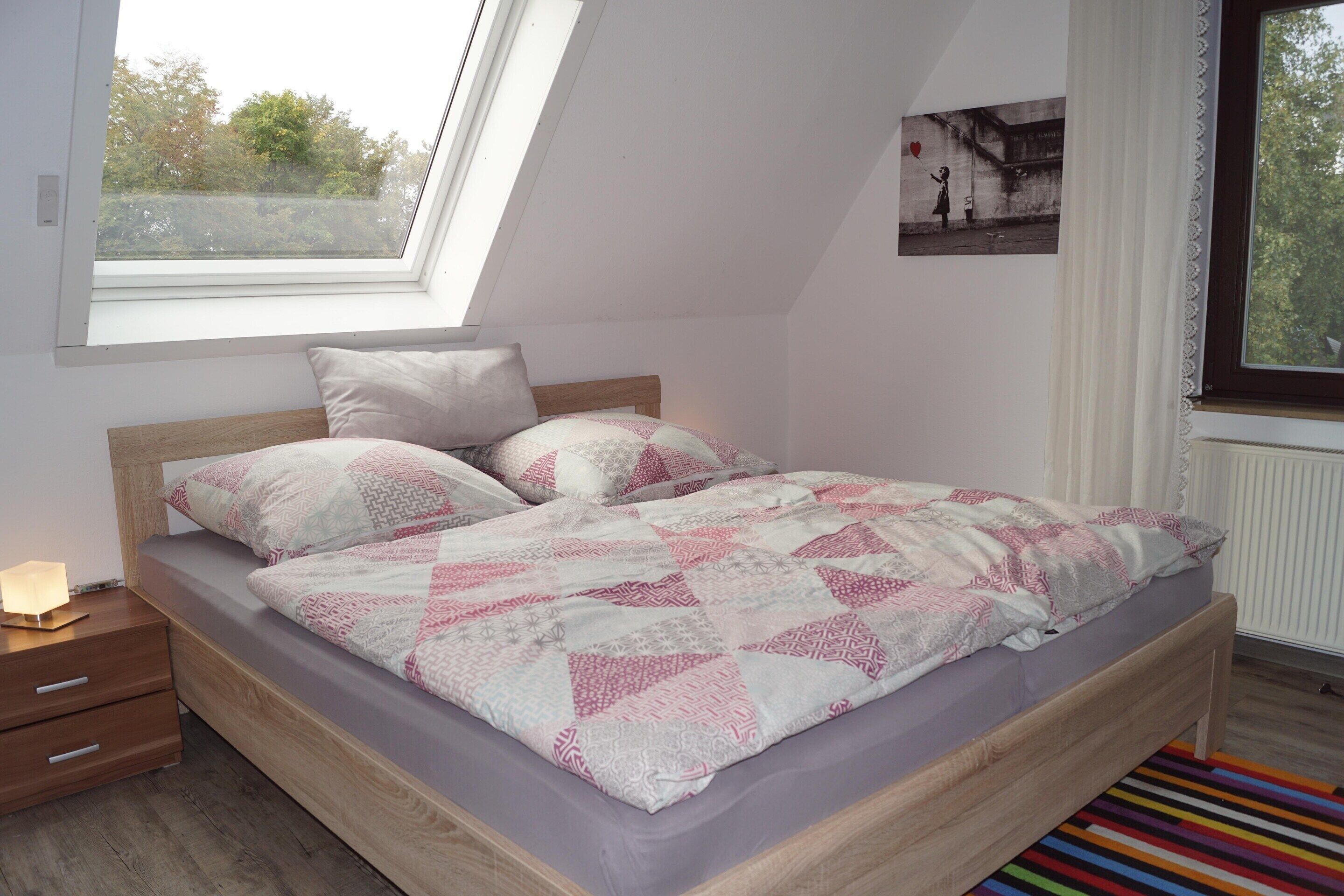 Una camera da letto con soffitto spiovente, un letto con copripiumino fantasia, un comodino con una lampada e una finestra vista alberi.