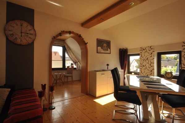 Une pièce avec un plancher en bois, une table à manger avec des chaises, une horloge au mur et un tableau encadré.
