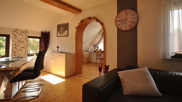 A modern living room with a black sofa, a dining table with chairs, a kitchen area, and a large wall clock. - Wohnung in Frankenberg fur Radfahrer (Frankenberg)