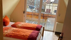 Een slaapkamer met een houten bed, beddengoed in oranje en rood, een raam met uitzicht op een balkon en gebouwen.