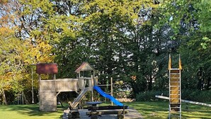 A wooden play structure with a slide and climbing area, surrounded by grass and trees. - Ferienhaus mit Terrasse im Skigebiet (Willingen)