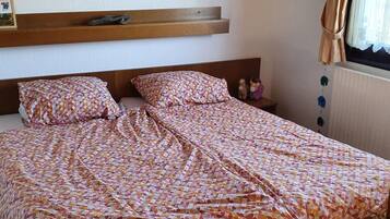 A bed with patterned bedding, a wooden headboard, a small bedside table with a lamp, and a wall-mounted shelf.