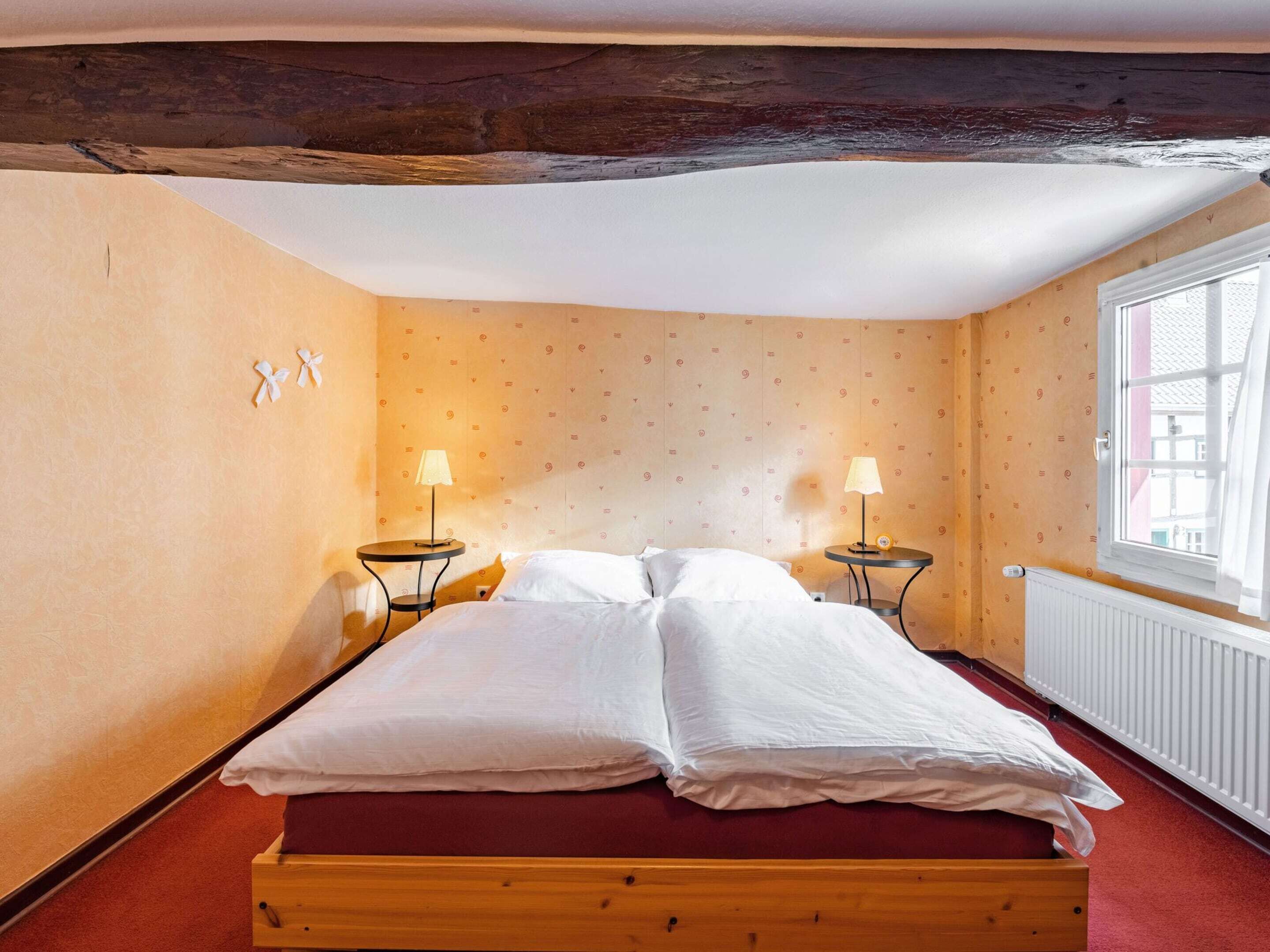Una habitación con cama de madera, dos mesitas de noche con lámparas, una ventana con vistas y papel tapiz estampado.