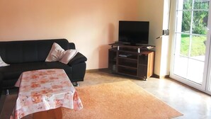 Ein Wohnzimmer mit einer schwarzen Couch, einem Fernseher auf einem Holztisch, einem Tisch mit einer Blumen Tischdecke und einem großen Fenster mit Blick auf Grünflächen.
