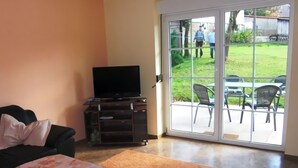 Ein Wohnzimmer mit einer Couch, einem Fernseher auf einem Holzboden, einem Tisch mit einer blumigen Tischdecke und einer Glastür zum Garten mit Stühlen und Rasen.