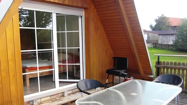 Eine Holzterrasse mit Glastisch und Stühlen, eine Glastür zum Schlafzimmer und ein kleiner Tisch mit Stuhl im Freien.