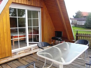 A wooden deck with a glass table and chairs, a sliding glass door leading to a bedroom, and a small outdoor table with a chair. - Holiday Home in Thale in the Harz Mountains (Thale)