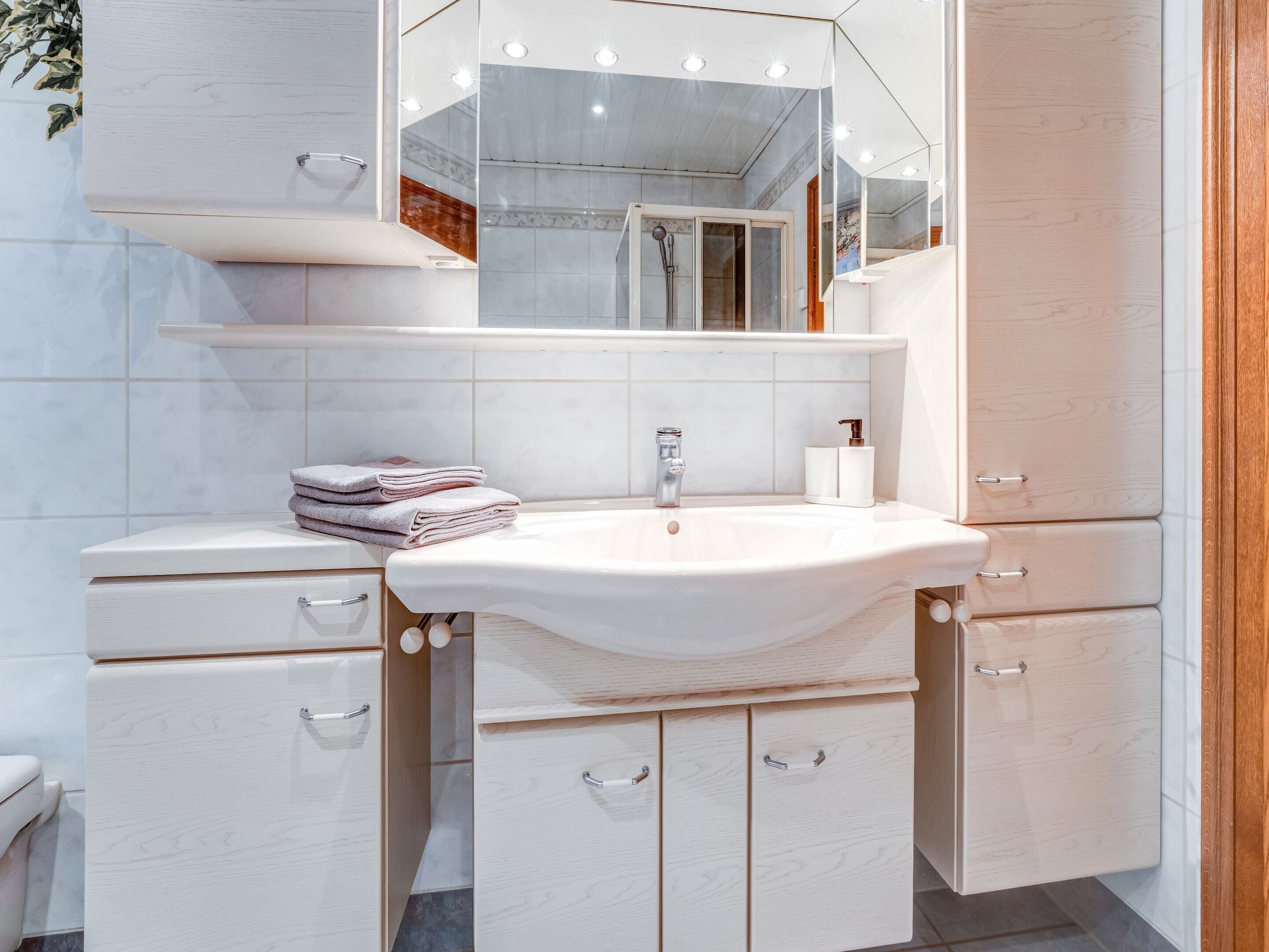 A modern bathroom with a white sink, vanity, and mirror. There are towels stacked on the vanity, and a shower door is visible in the background.