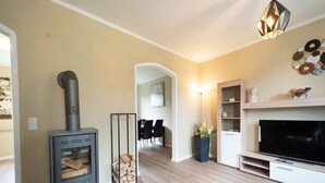 A modern living room with a wood-burning stove, a flat-screen TV, and a dining area in the background. - Holiday Home With Garden (Rechenberg-Bienenmuehle)