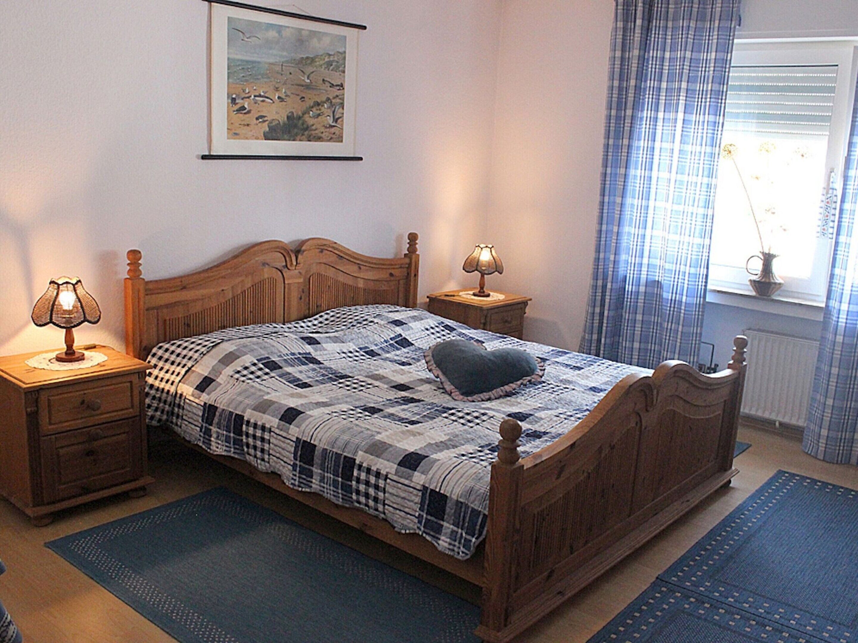 A bedroom with a wooden bed, two bedside tables with lamps, a blue and white checkered bedspread, a heart-shaped pillow, a painting on the wall, and a window with blue curtains.
