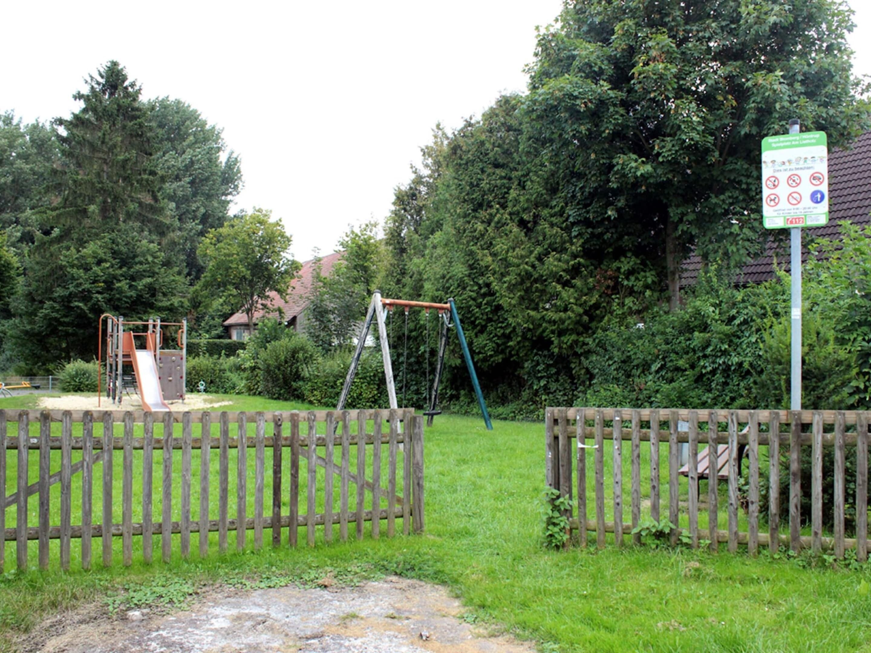 A playground with a slide and swings, surrounded by a wooden fence and greenery.