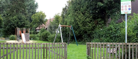 A playground with a slide and swings, surrounded by a wooden fence and greenery.