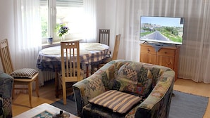 A living room with a dining table, chairs, a sofa, a television, and a window with curtains. - Wohnung im Teutoburger Wald (Blomberg)