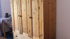 A wooden wardrobe with multiple doors and drawers, a small table with a cloth and a kettle, and a chair. - Wohnung im Teutoburger Wald (Blomberg)