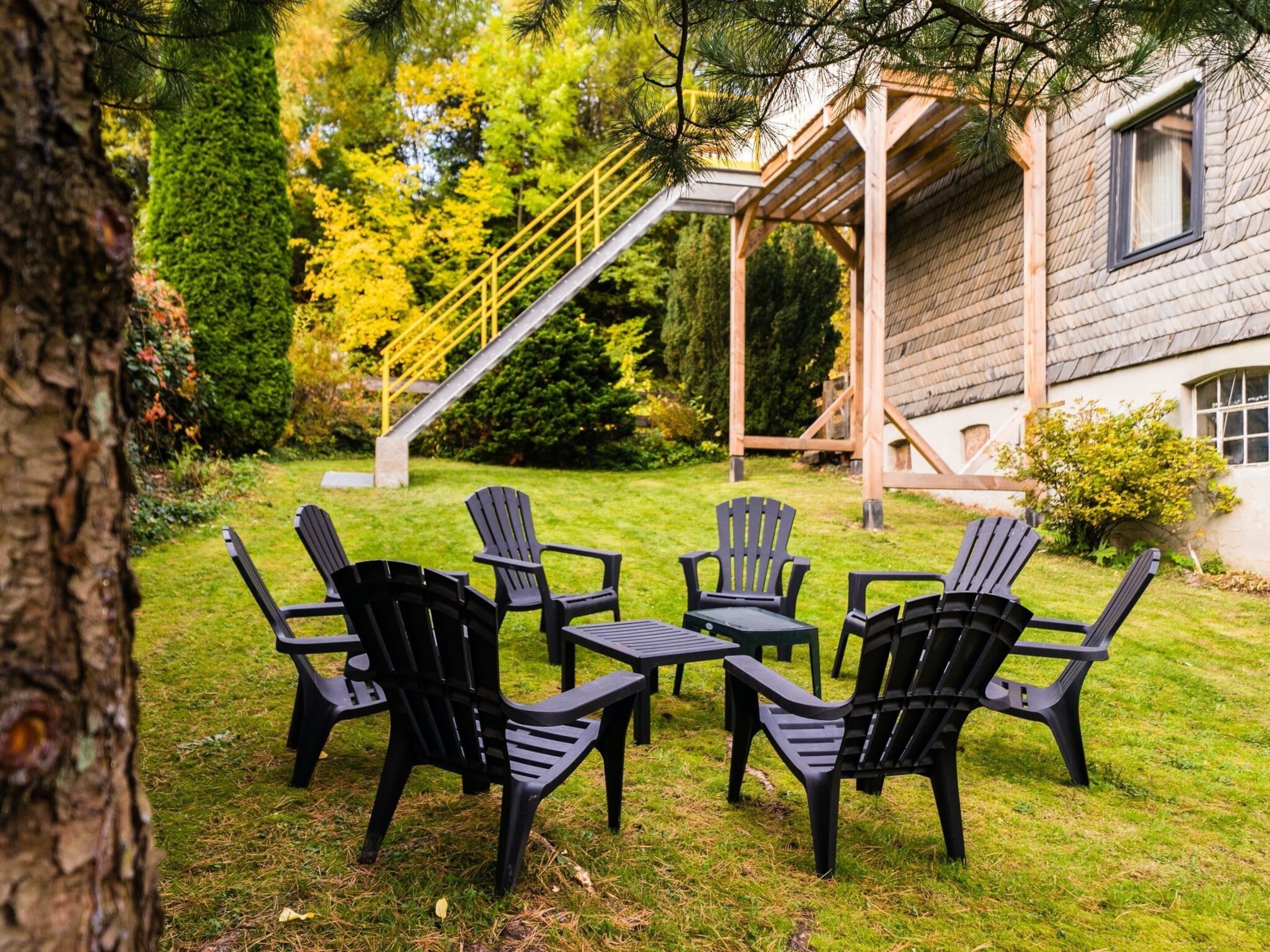 A patio with six black Adirondack chairs arranged around a small table, a wooden pergola, and a staircase leading to a house with a window.