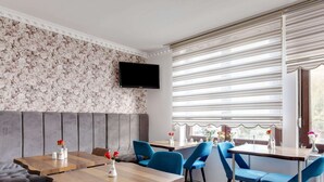 A modern dining area with a sofa, wooden tables, and blue chairs. A flat-screen TV is mounted on the wall, and there are windows with blinds. - Lakeside Base in Oberhausen (Oberhausen)