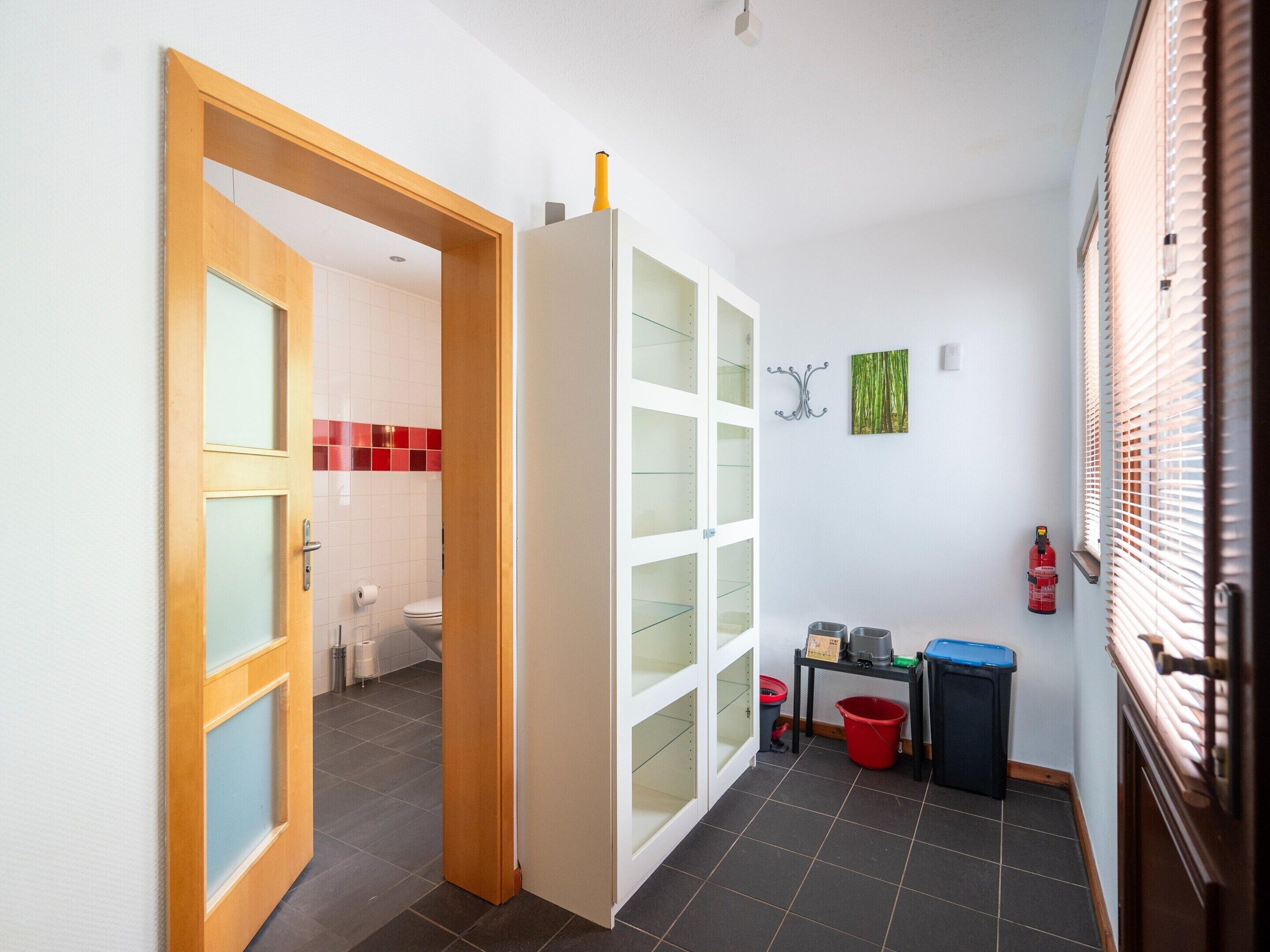 A hallway with a wooden door leading to a bathroom, a white cabinet with glass doors, a small table with a red bucket, and a fire extinguisher on the wall.
