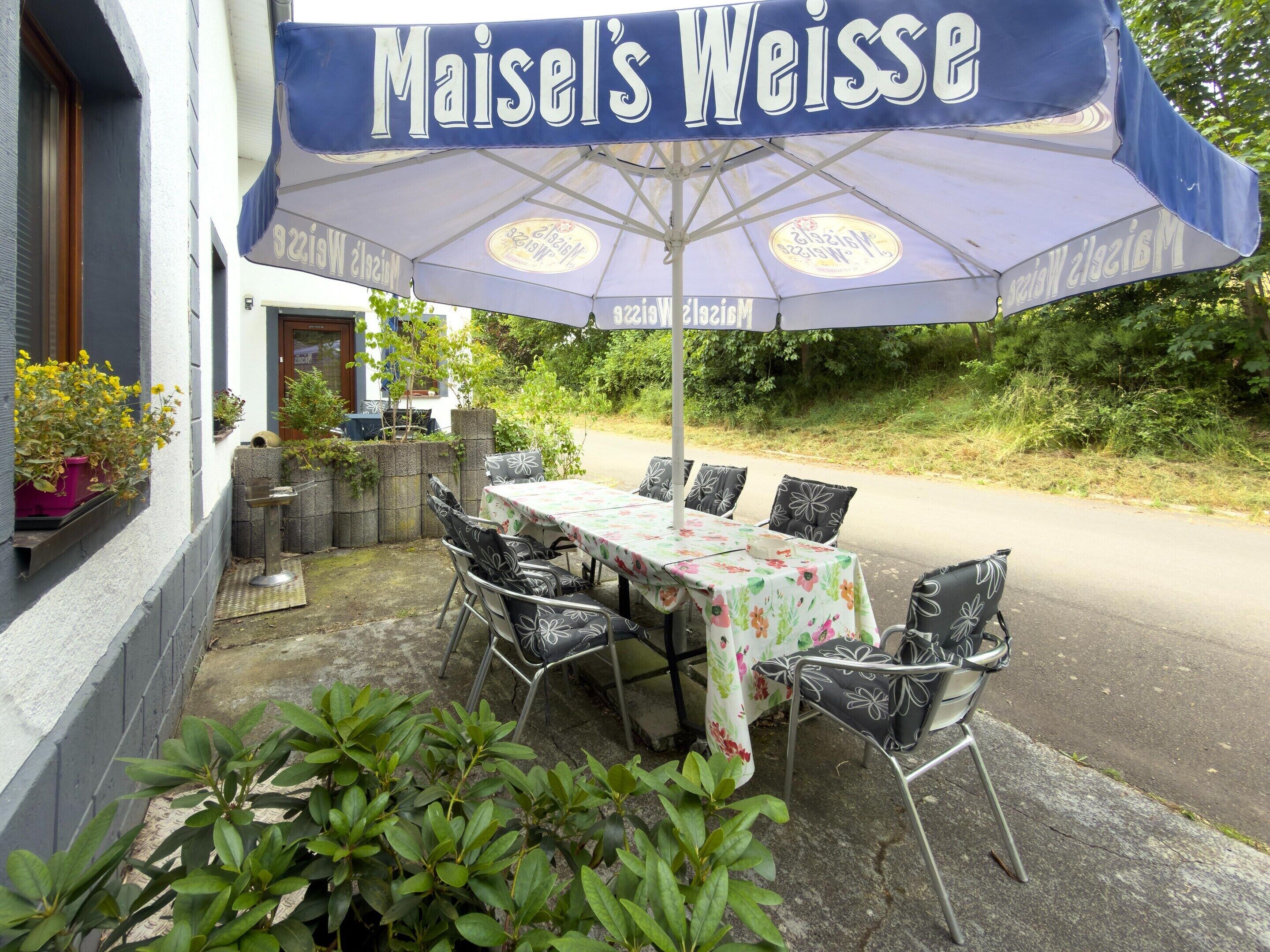 Un área de comedor al aire libre con mesa y sillas bajo un gran paraguas que dice 'Maisel's Weisse'.