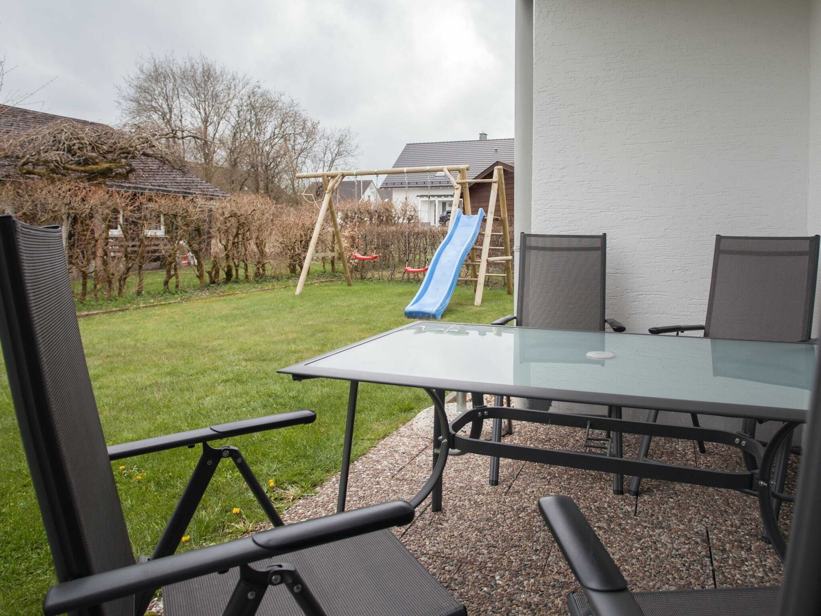 Een terras met een glazen tafel en stoelen, een speelstructuur voor kinderen en een gazon.