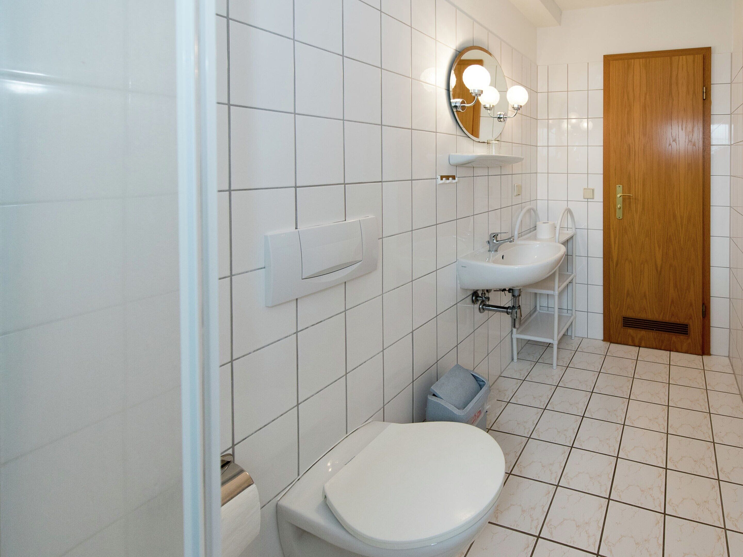 A bathroom with white tiles, a toilet, a sink, a mirror with lights, and a wooden door.