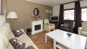 Een woonkamer met een witte bank, een witte salontafel, een open haard, een televisie en een raam met gordijnen.