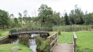 Een houten brug over een beekje in een park met grasrijke oevers en bomen op de achtergrond.