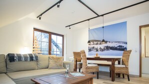 A living room with a sofa, coffee table, and wicker chairs around a wooden dining table. A large window with a view of a snowy landscape is visible. - Apartment With Balcony in Neuastenberg (Winterberg)