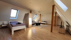 A spacious loft bedroom with a high ceiling, wooden beams, a large bed, a desk, and a skylight.