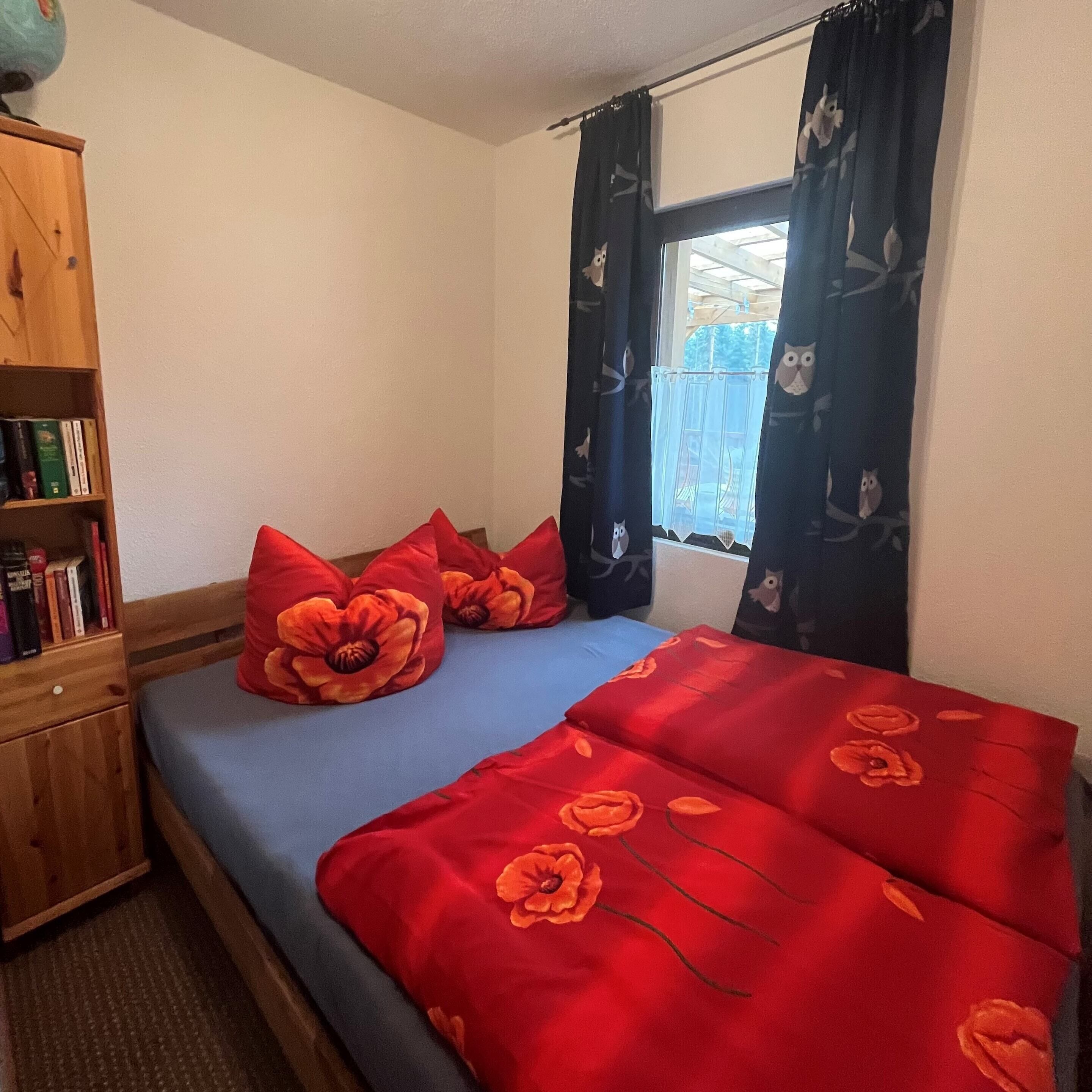 A bedroom with a bed, red floral pillows, a blue bedspread, a wooden bookshelf, and a window with curtains.