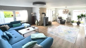 A modern living room with a blue sofa, a coffee table, and a rug. There is a fireplace, a dining area, and a kitchen visible. - Wellness & Comfort Retreat (Fehmarn)