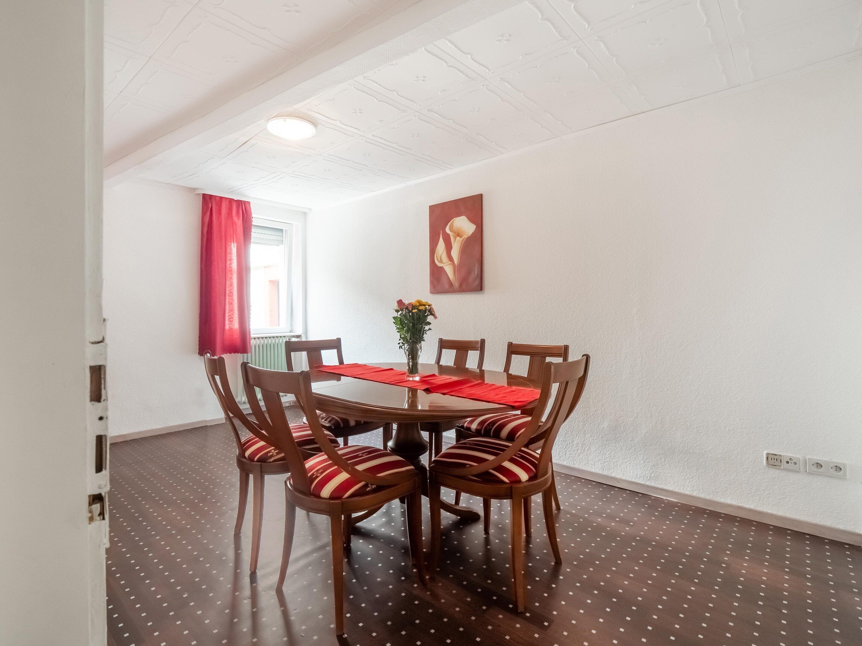 A dining area with a round table, six chairs, a vase with flowers, and a painting on the wall.