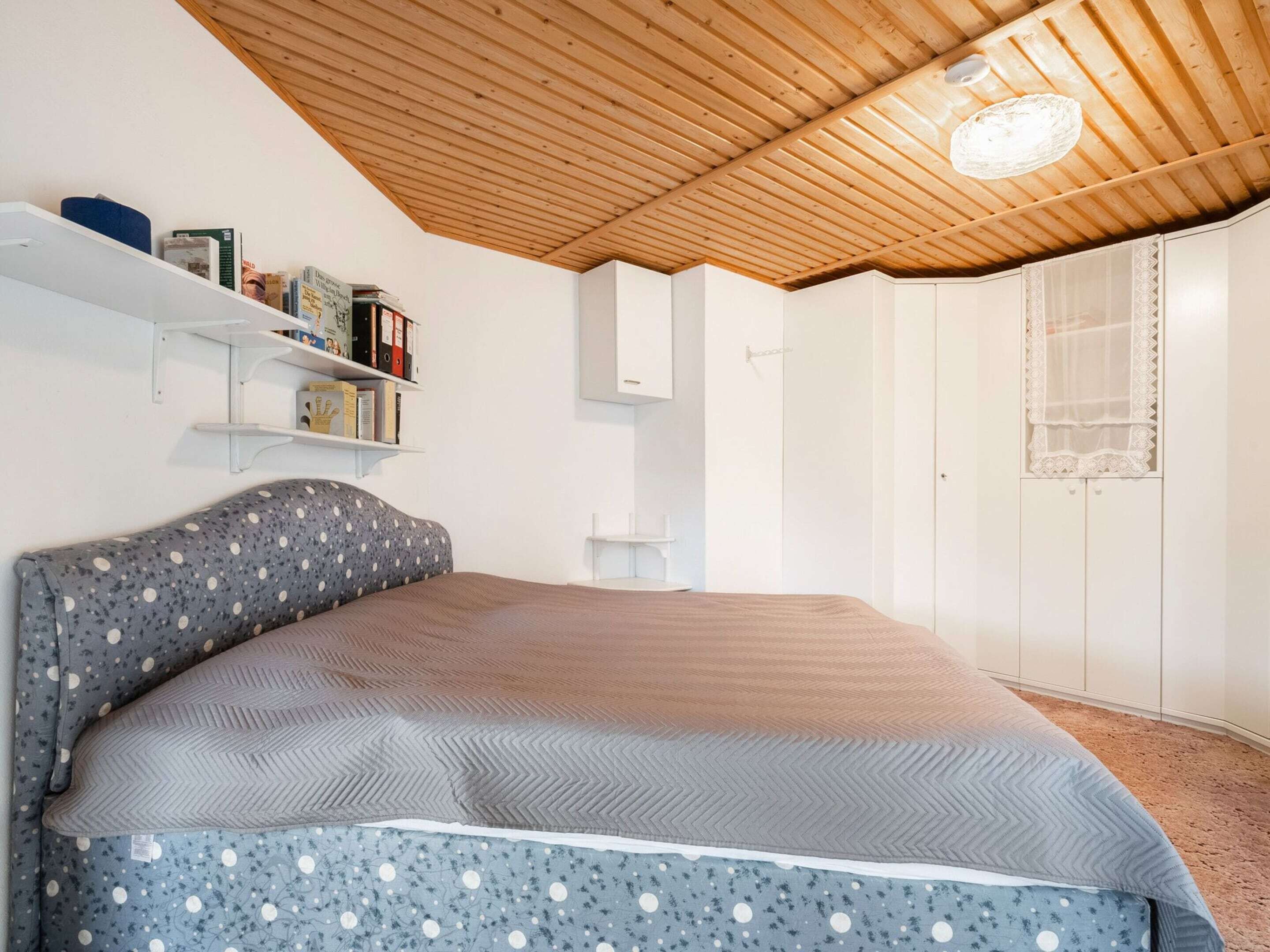 Ein Schlafzimmer mit Holzbalkendecke, einem Bett mit Kissenüberwurf in Punkten und Zickzackmuster, weißen Wänden und einem weißen Kleiderschrank mit Spitzenverzierung.