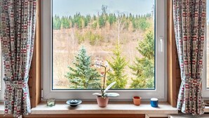 A window with a view of a forested area, a potted plant, and a small table with a candle and a cup. - Ferienhaus in Harzgerode (Harzgerode)