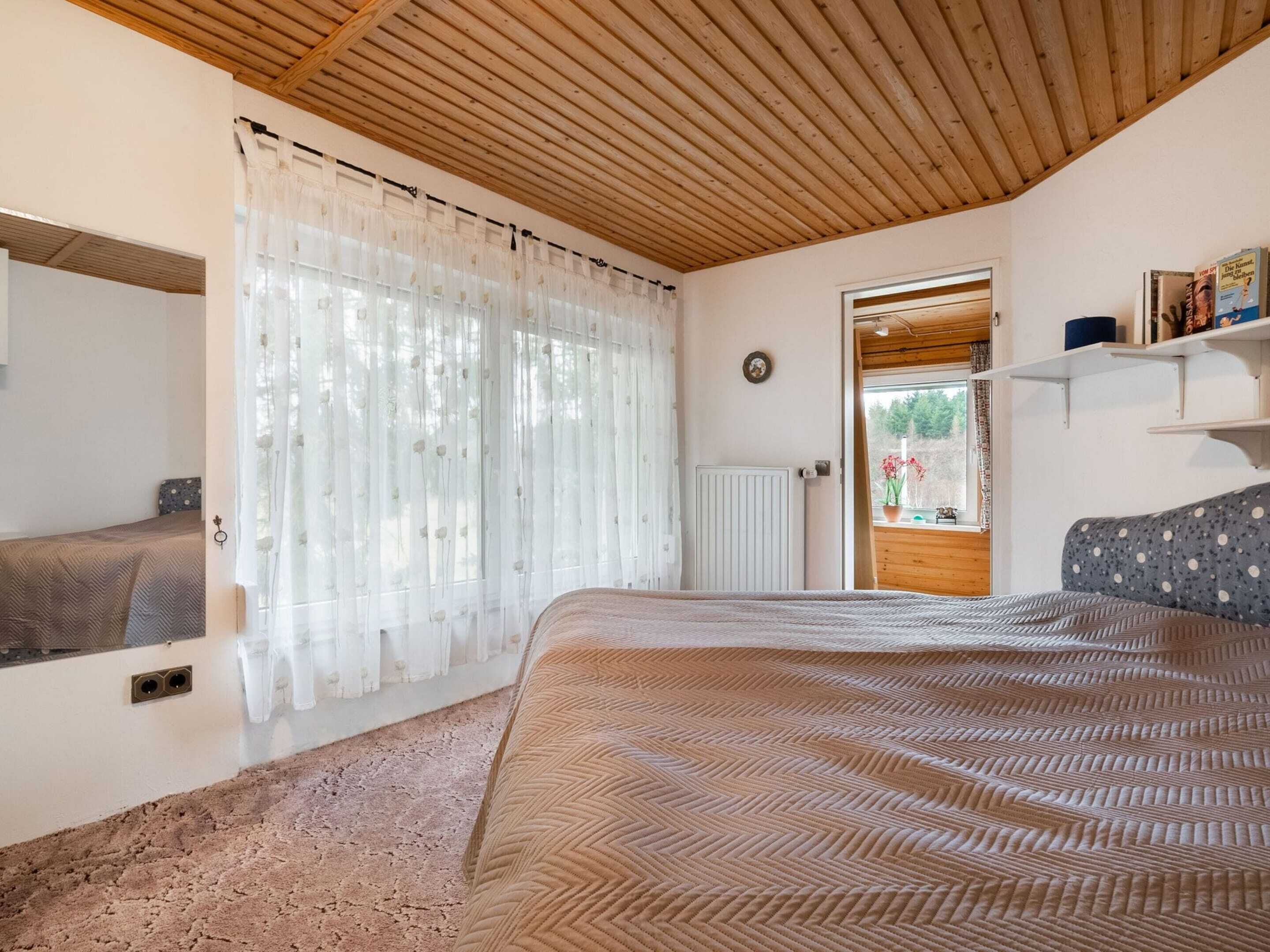 A bedroom with a bed, a window with sheer curtains, a radiator, and a wooden ceiling.