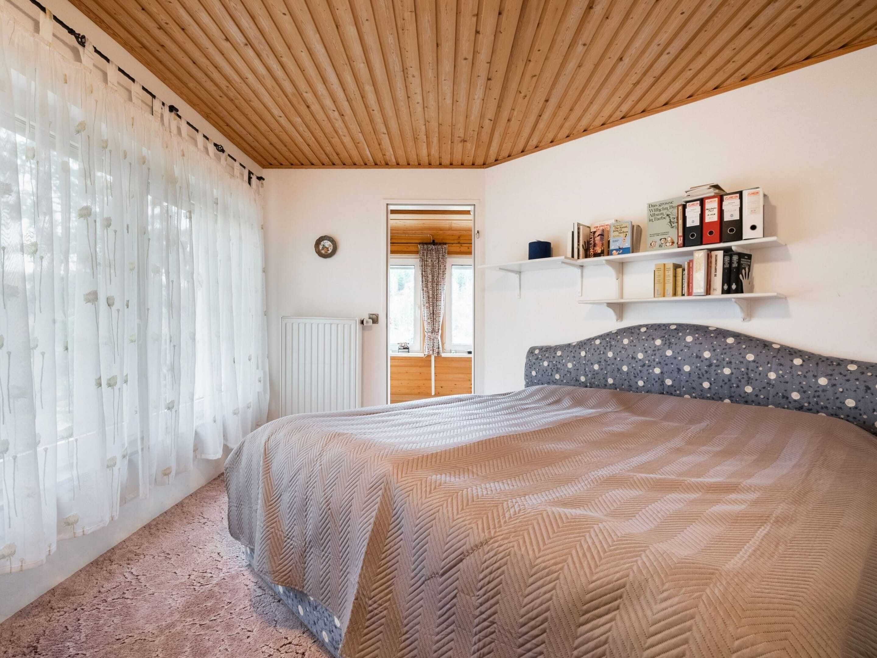 A bedroom with a wooden ceiling, a bed with a patterned headboard, a radiator, and a bookshelf with books and binders.
