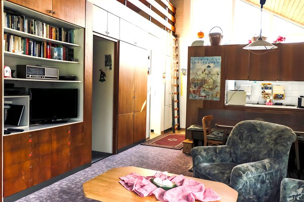 A compact living space with a bookshelf, a television, a dining area, and a chair. - Nordsee-ferienhaus mit Terrasse (Wangerland)