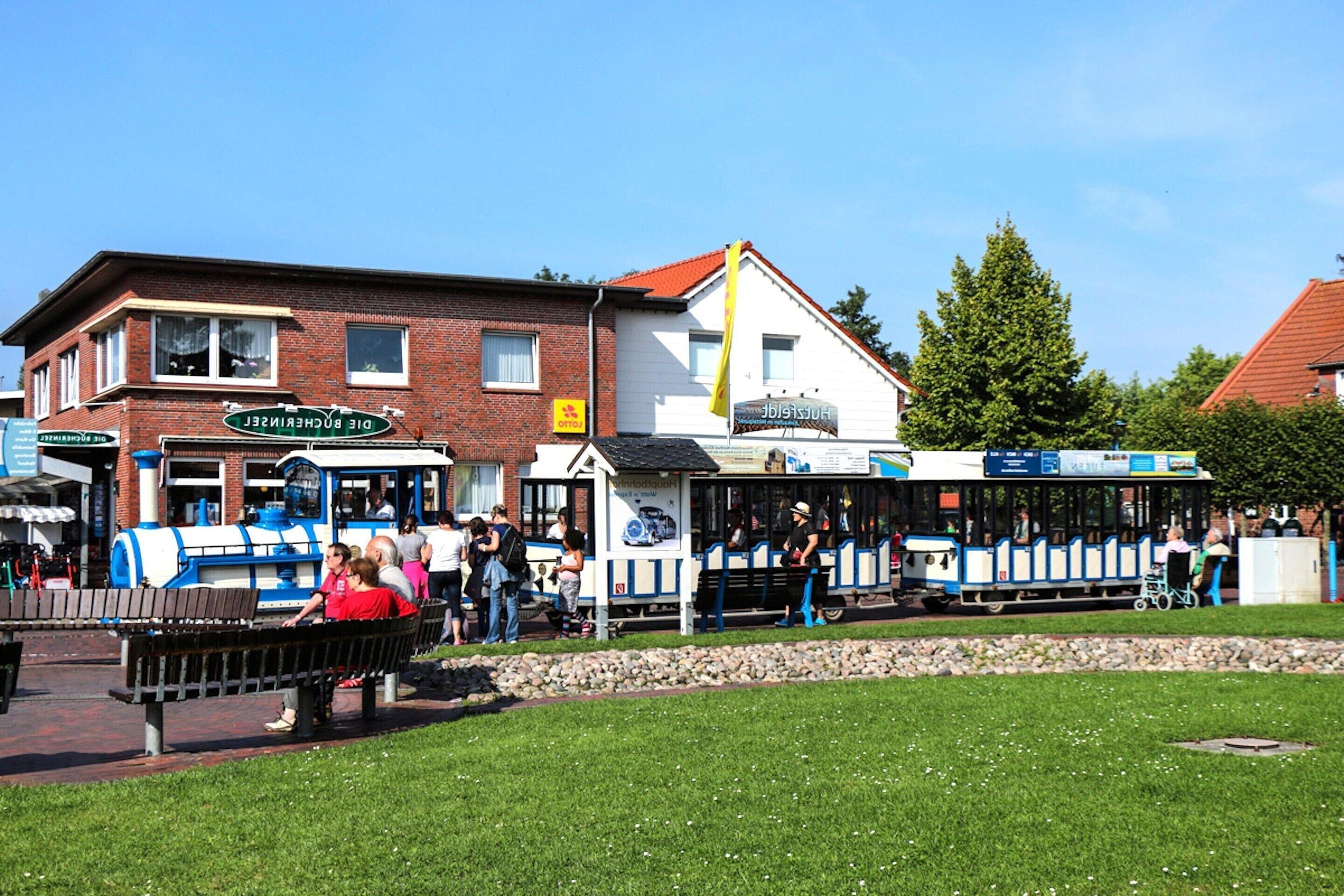 A train station with a blue and white train, a brick building with a sign 'DIE BÜCHNERSTELLE', and a grassy area with benches.