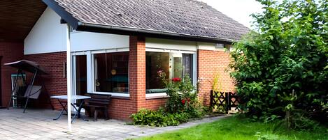 A house with a brick exterior, a tiled roof, a patio with a table and chairs, and a garden with a fence.