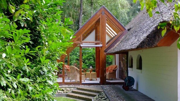 A wooden gazebo with wicker furniture, a thatched-roof structure, and a white building with a window.