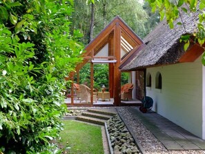 A wooden gazebo with wicker furniture, a thatched-roof structure, and a white building with a window. - Holiday Home in Uelsen Near Bronzezeithof (Uelsen)