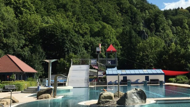 A swimming pool with a water slide, surrounded by a red-roofed building and a mountain backdrop. - Grosszugiger Komfort, Rubeland (Oberharz am Brocken)