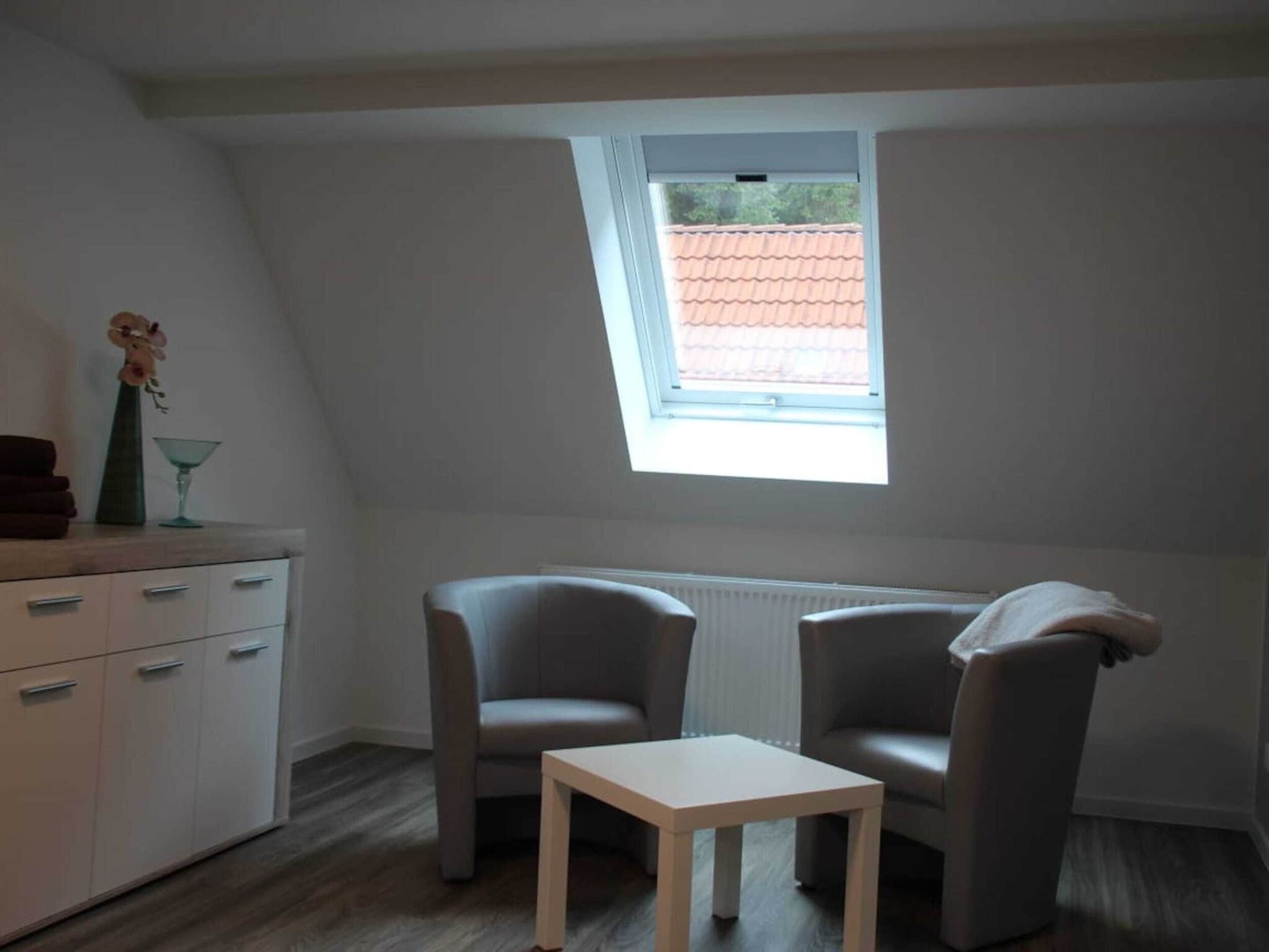 A small, well-lit room with a sloped ceiling, a skylight, two grey chairs, a small white table, a white cabinet with drawers, and a vase with flowers on the cabinet.