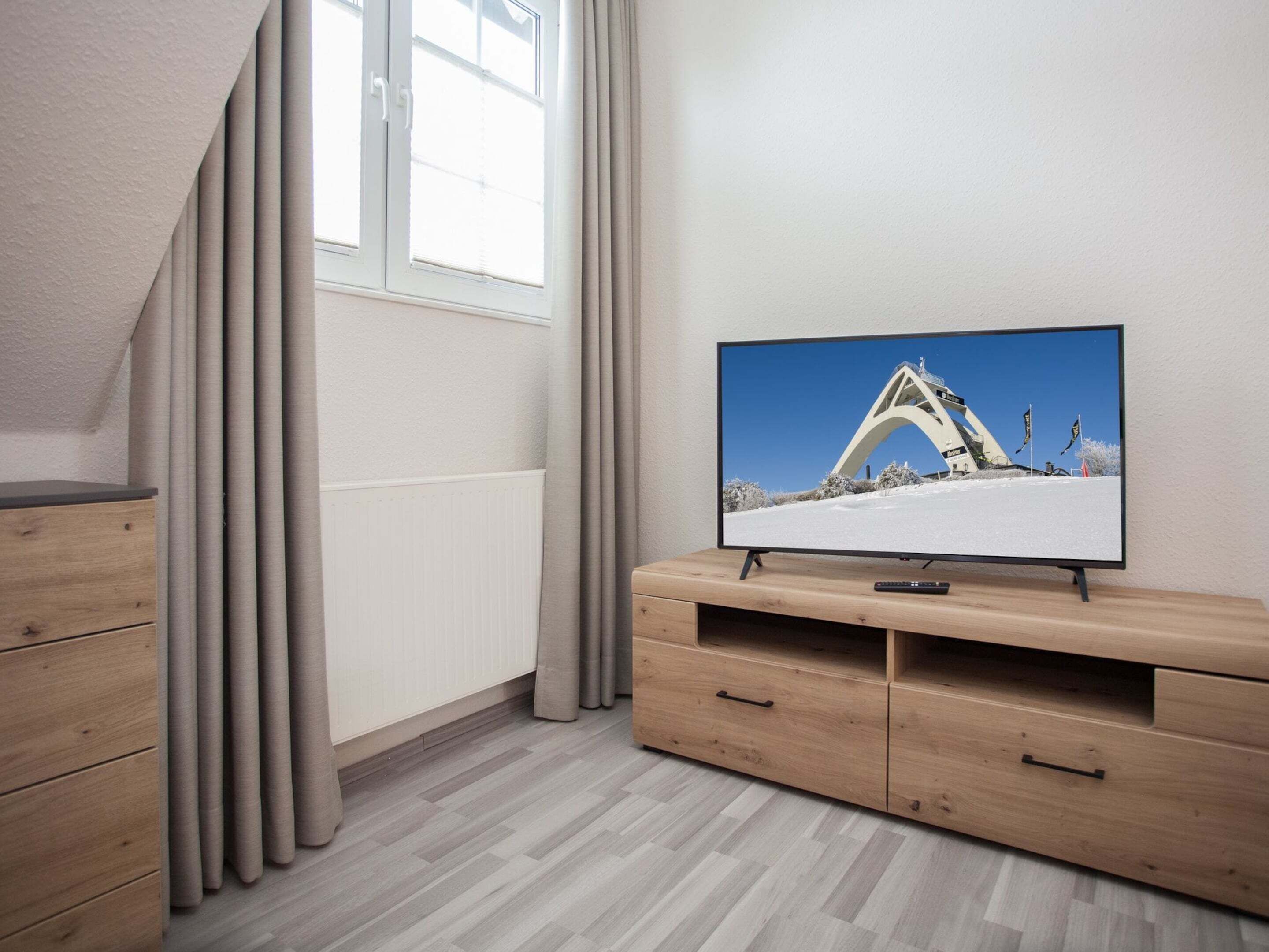 Moderni huone, jossa on televisio puisen jalustan päällä, ikkuna verhoilla ja patteri.