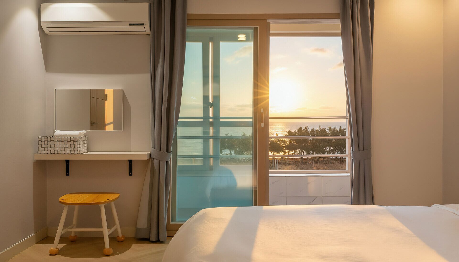Habitación de hotel con cama, un taburete pequeño, un espejo, una caja de almacenamiento y vistas a un puente y al atardecer.