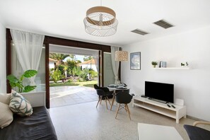 Een moderne woonkamer met een flatscreen televisie, een eethoek met een tafel en stoelen, en uitzicht op een tuin door een open deur.