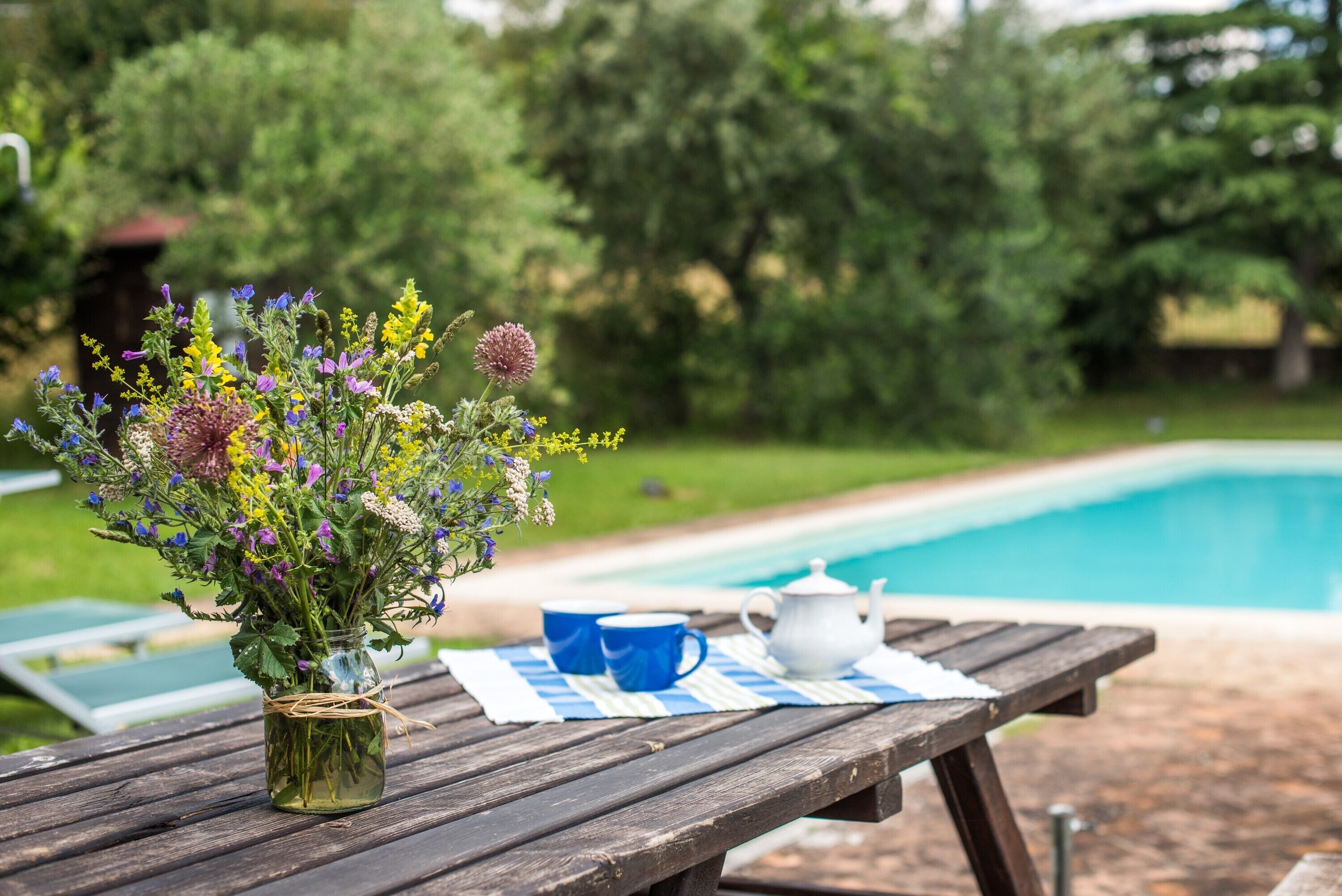 A wooden picnic table with a vase of flowers, two blue cups, and a white teapot.