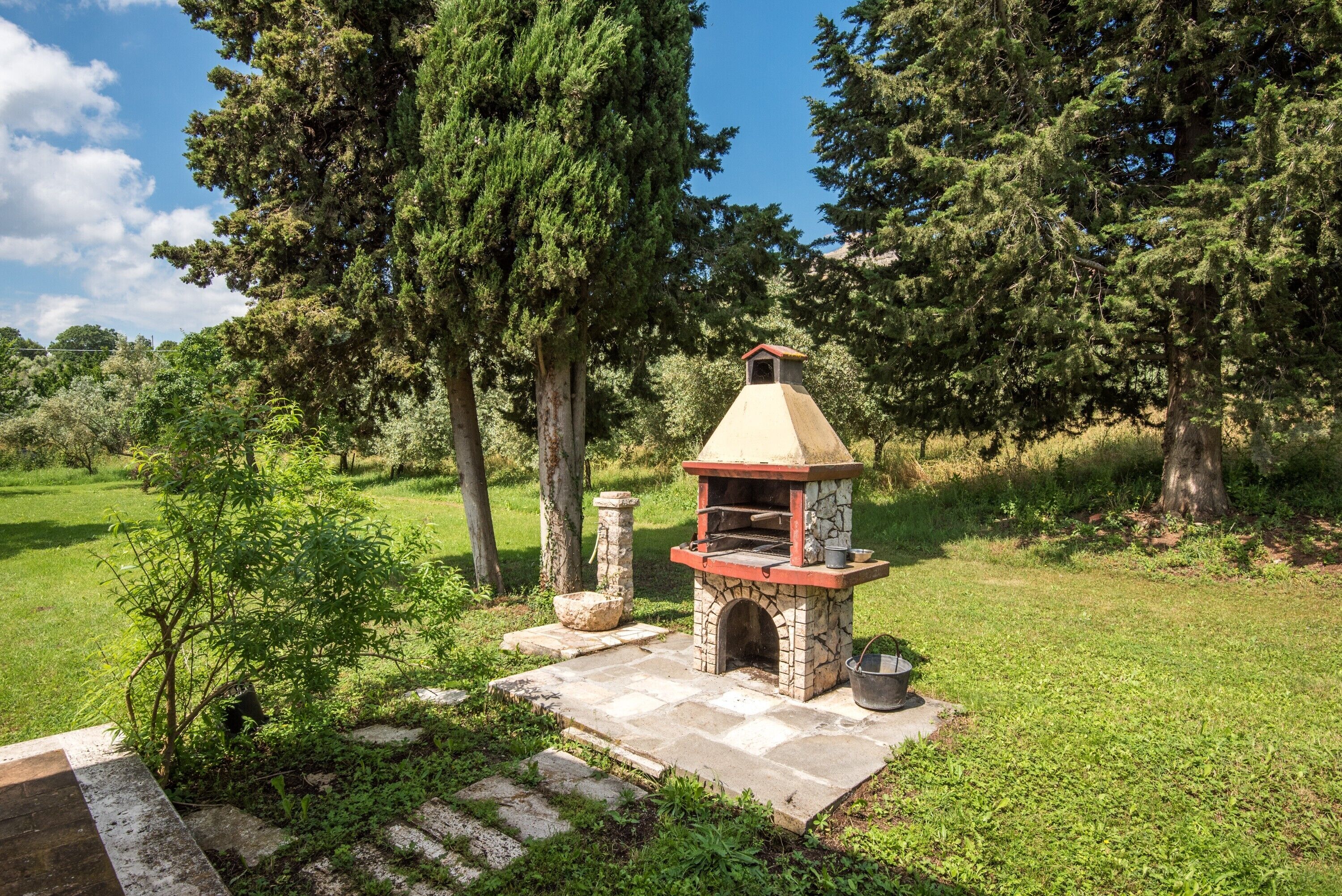 A stone-built outdoor oven with a chimney, surrounded by a grassy area and trees.