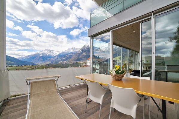 A balcony with a wooden table and chairs, a wicker basket with flowers, and a mountain view.